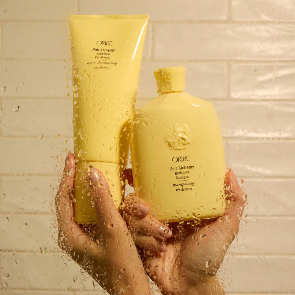 Oribe Hair Alchemy Resilience Shampoo and Conditioner held in hands under shower water for strengthening fragile hair