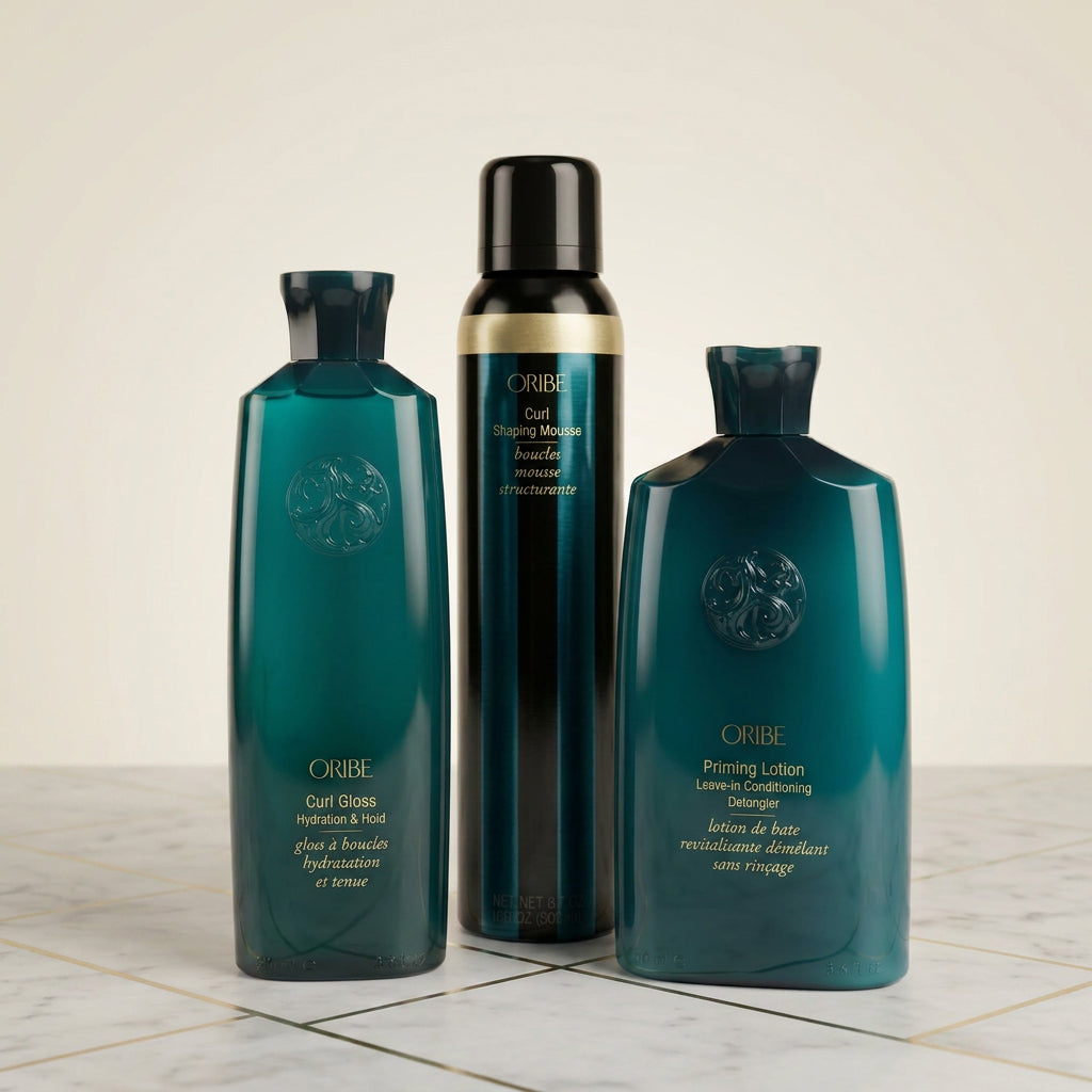 Three ORIBE haircare products in dark teal and black bottles with gold lettering, arranged on a light marble surface against a warm beige background.
