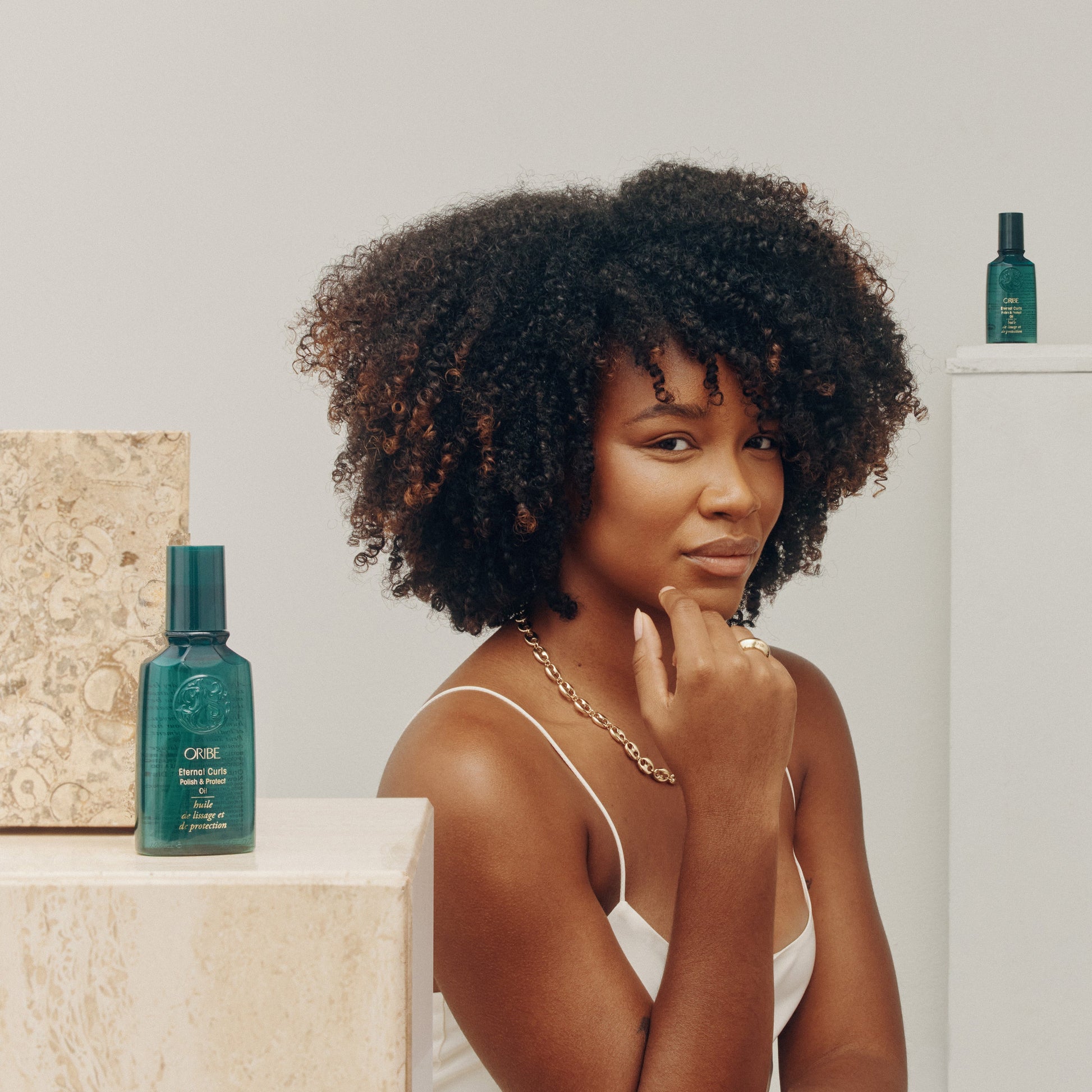 Woman with curly hair sitting next to a Oribe Eternal curls Polish and Protect oil on a white background