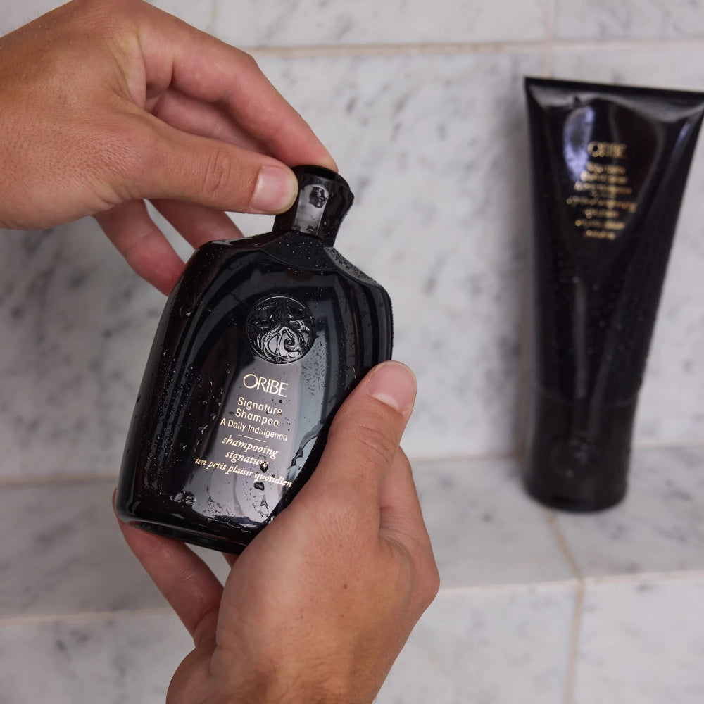 Close-up of hands opening an Oribe Signature Shampoo bottle during a luxury hair washing routine.